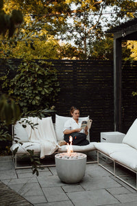 Woman reading a book on a patio with a fire pit and white outdoor furniture.