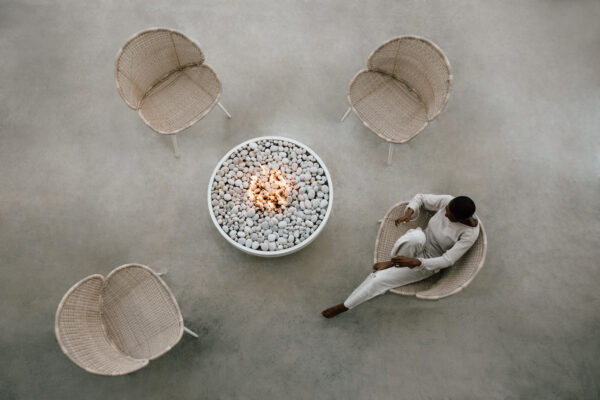 Person sitting on a chair around a fire pit with four chairs arranged around it on a concrete floor.
