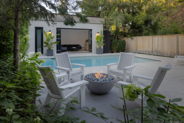 Outdoor patio with fire pit, chairs, and pool area surrounded by greenery